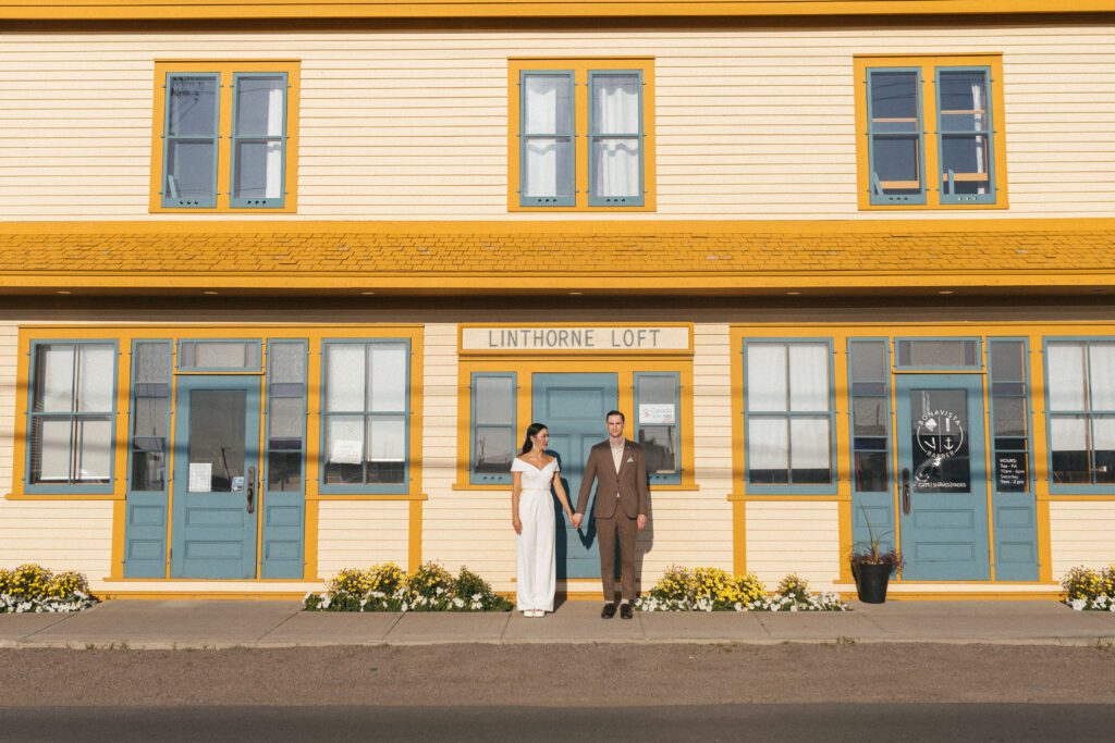 bride and groom hold hands in front of vibrant yellow and blue building in Bonavista, Newfoundland