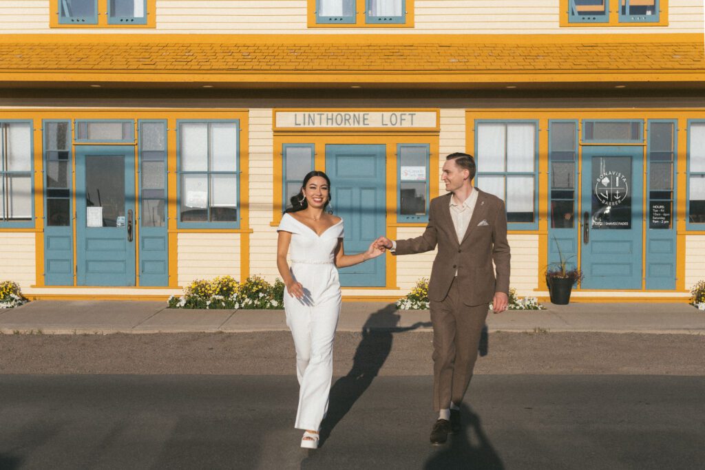 bride and groom laugh as the run in front of vibrant yellow and blue building in Bonavista, Newfoundland