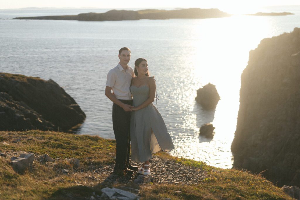 bride and groom hold each other on a rocky cliff with the sun glinting on the ocean behind them