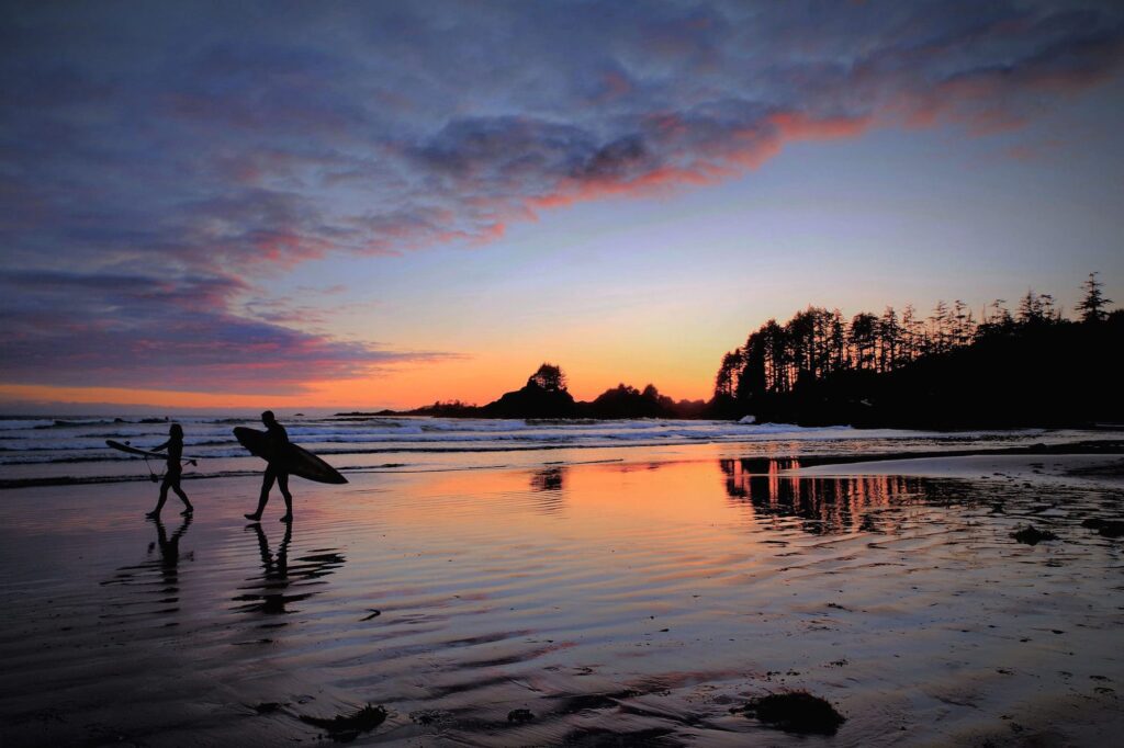 two silhouetted figures carry surf boards along a sandy beach at sunrise
