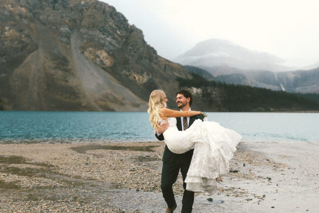 groom carries bride across a river and grins at her