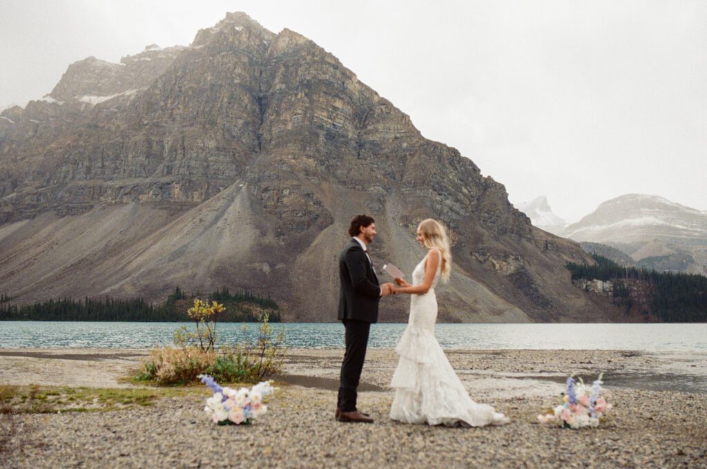 bride and groom read each other vows on the shore of Bow Lake with mountains behind them 