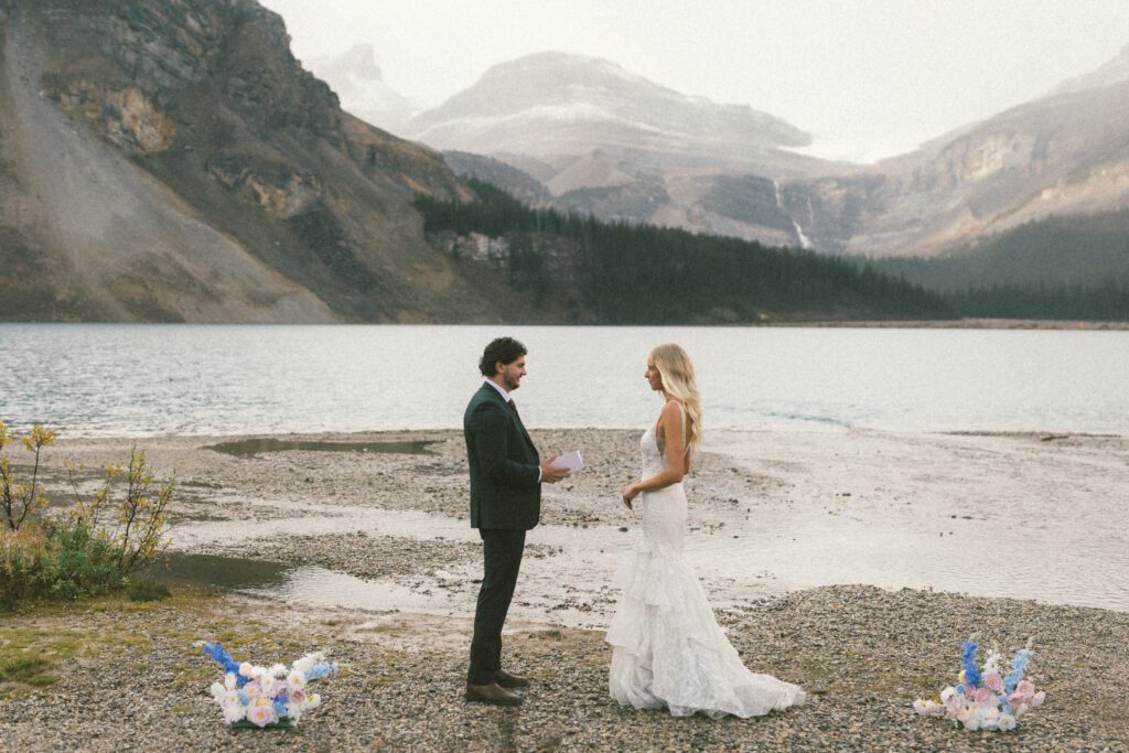 bride and groom read each other vows on the shore of Bow Lake with mountains behind them 