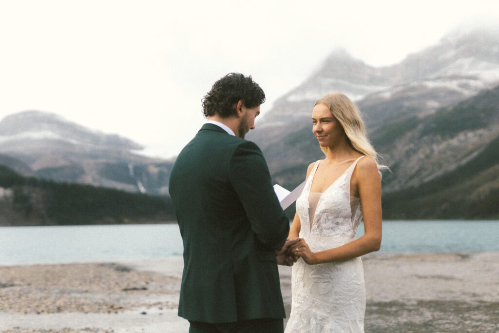 bride smiles at groom and holds his hand as he reads his vows beside a mountain lake in Banff