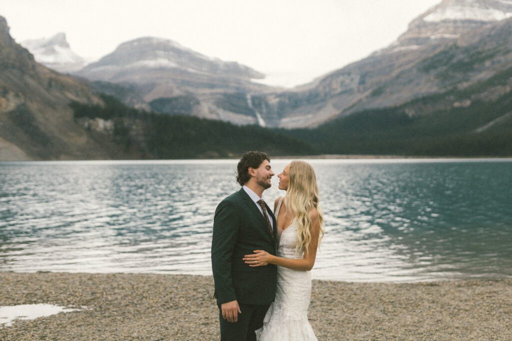 bride and groom hold each other and smile beside a glacier lake with mountains behind 