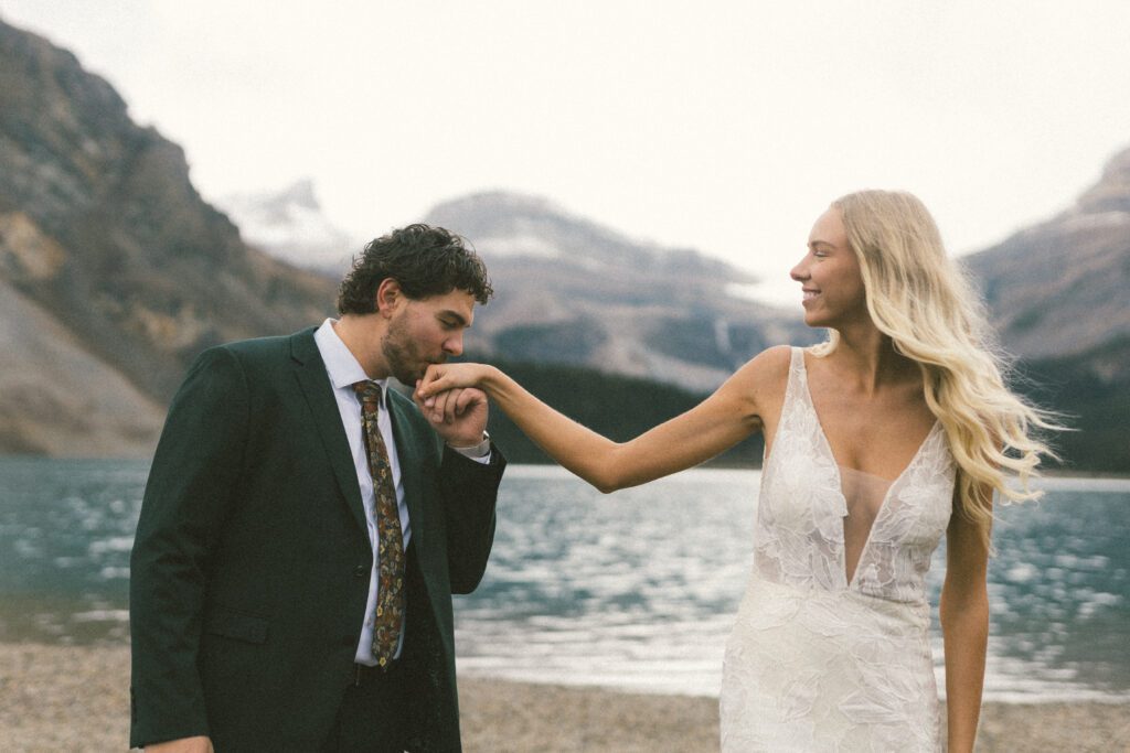 groom kisses bride's hand and she laughs by a mountain lake