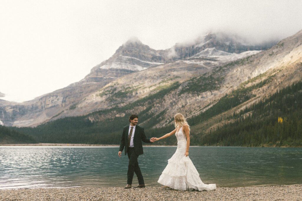 bride and groom hold hands and walk along the shore of a mountain lake