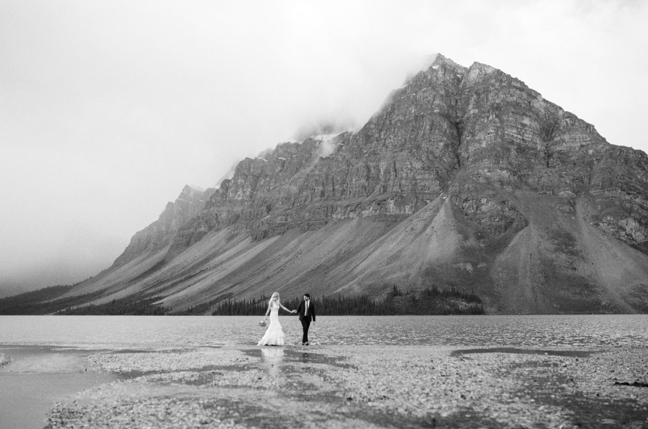bride and groom hold hands and walk along the shoreline of a mountain lake in Banff