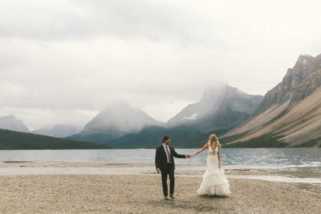 bride and groom wander along a lakeshore in Banff holding hands