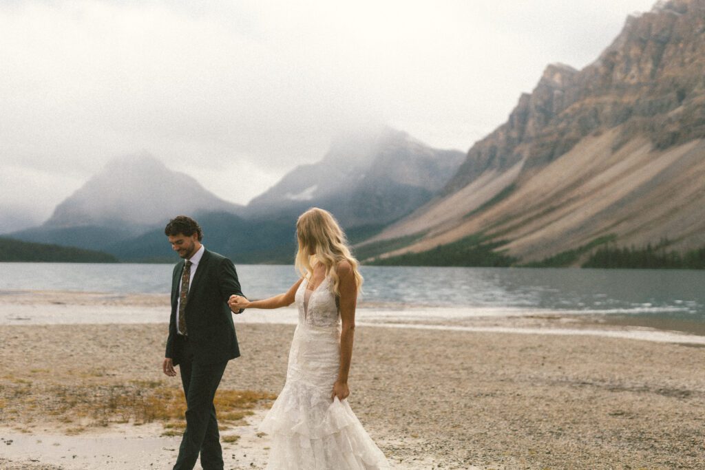 bride and groom walk along a sandy lake shore with mountains shrouded in mist behind them