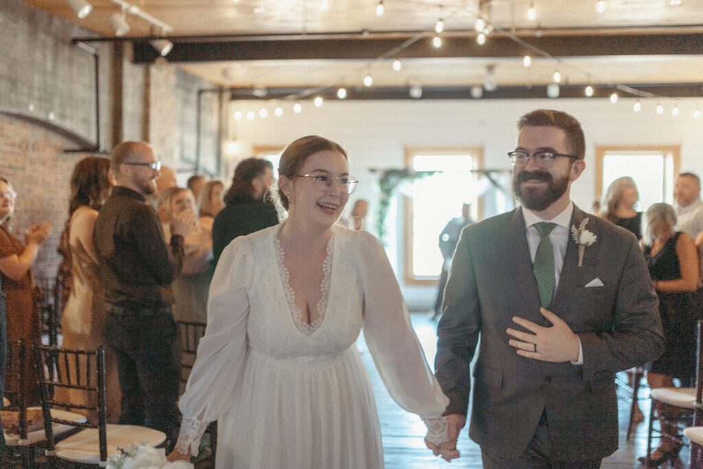 bride and groom laugh as they walk down the aisle in a bright space with wooden beams and exposed brick