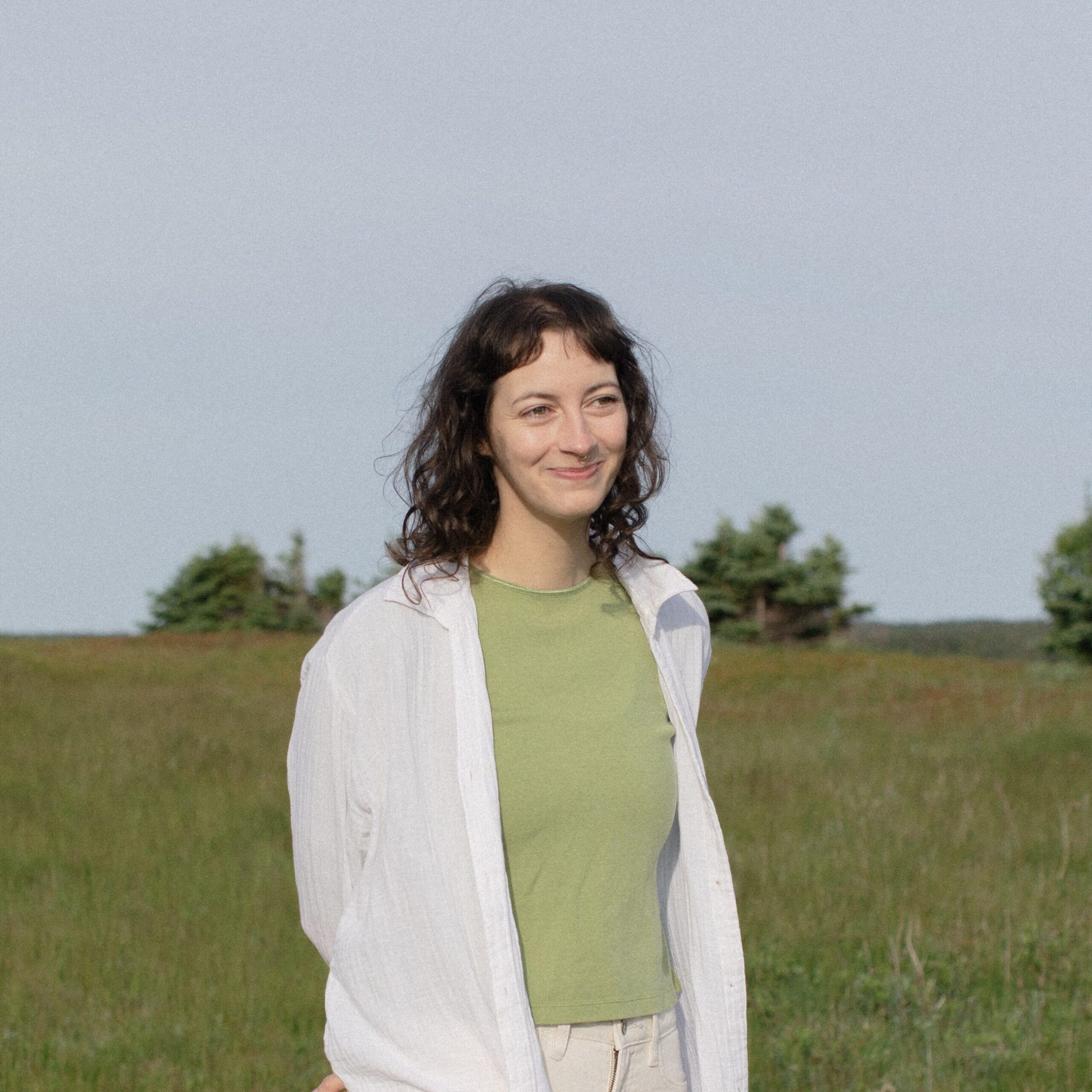 a non-binary person with dark curly hair walks through a meadow of tall grass, smiling and looking to the side