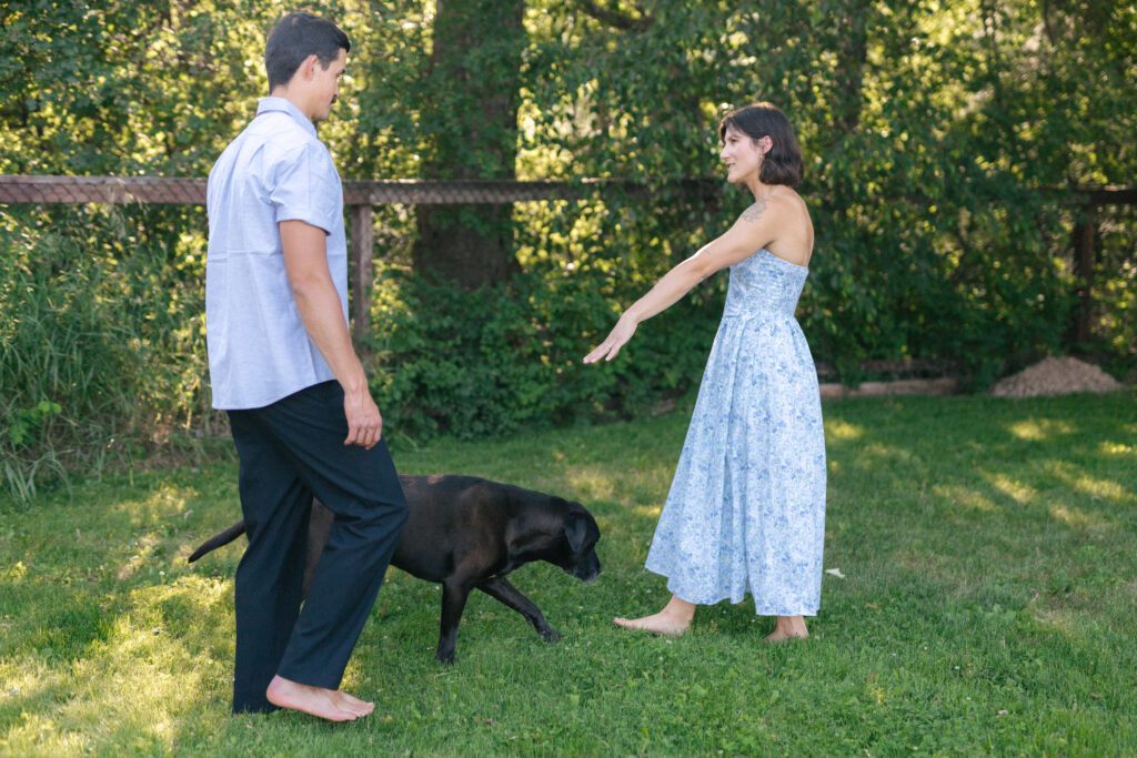 woman in a blue dress reaches out a hand to her husband in a yard surrounded by trees as their dog walks past