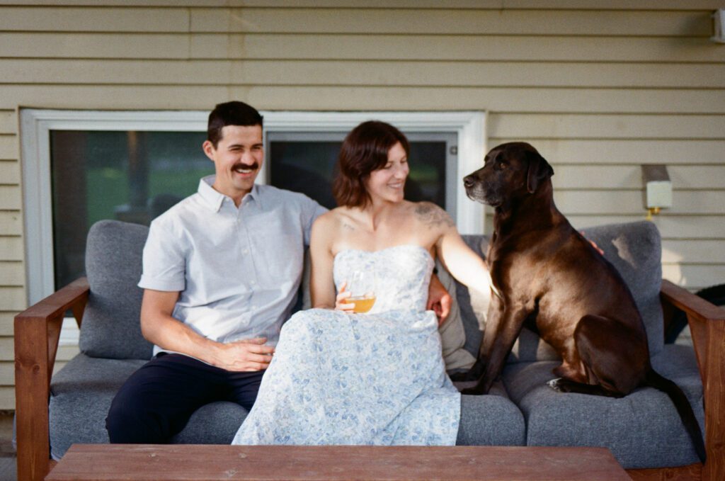 man and woman sit on an outdoor couch laughing and petting a brown dog