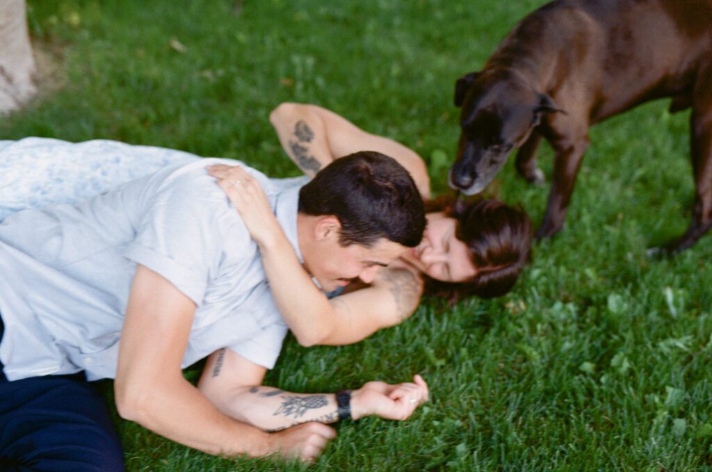couple wrestle playfully in the gras while their dog licks their faces