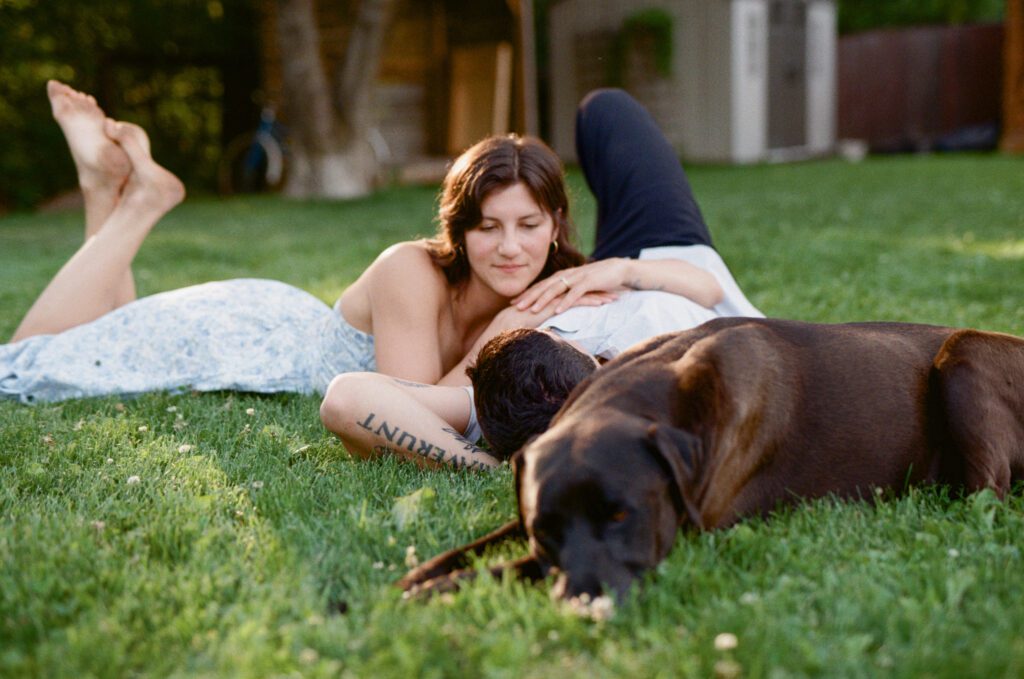 woman lies on man's chest in the grass, with their dog lying beside them
