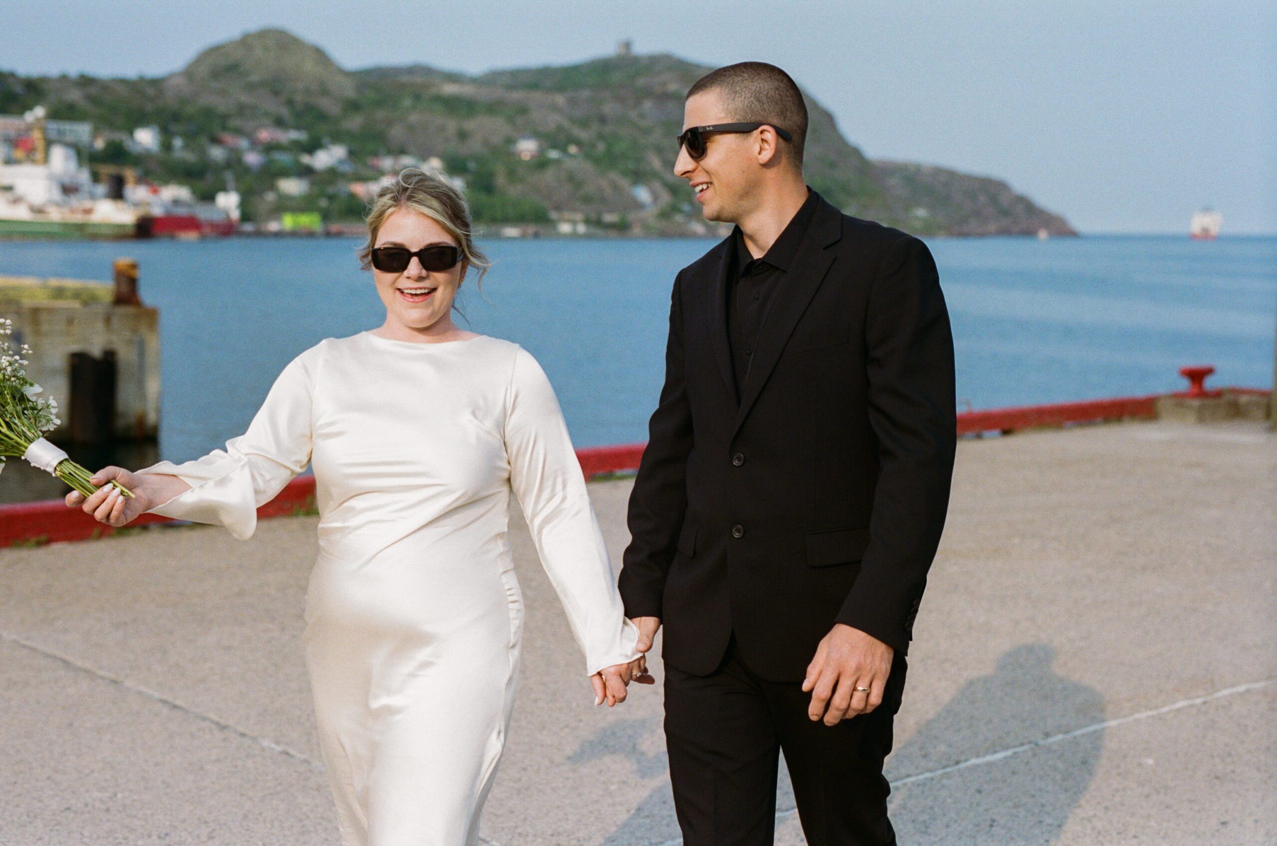 35 mm film photo of a bride and groom in sunglasses laughing as they walk along the harbour in St. John's, Newfoundland
