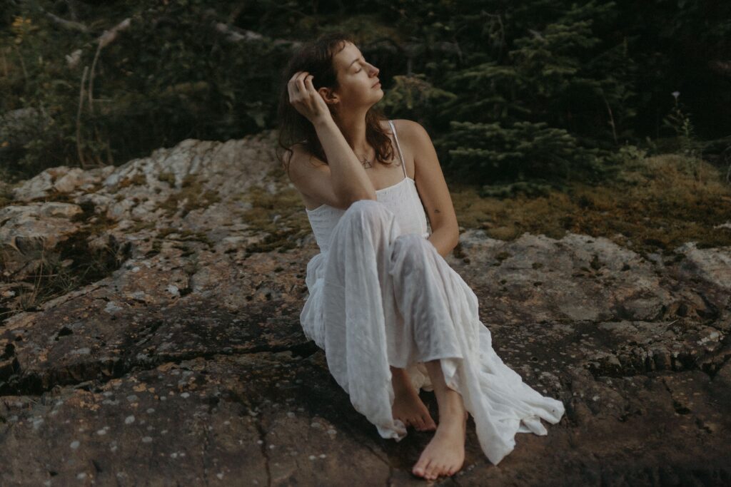 woman in a long white dress sits on a rock with their eyes closed and a serene expression during Self Love Session in BC